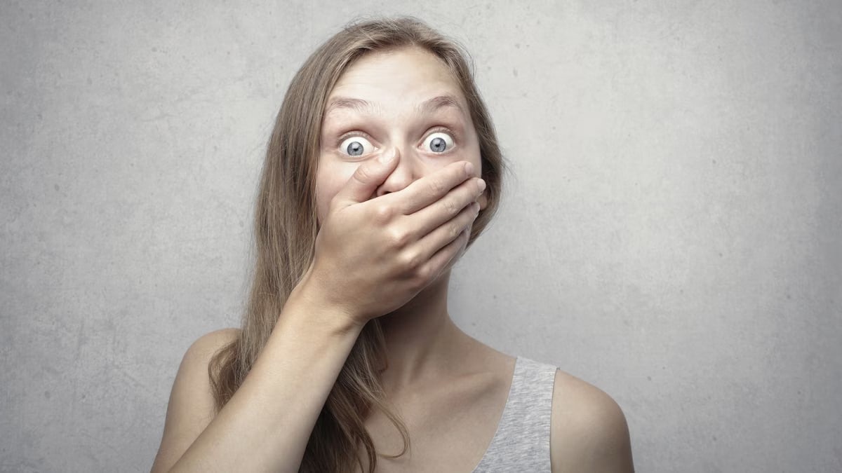 A startled woman covers her mouth expressing surprise against a neutral background.